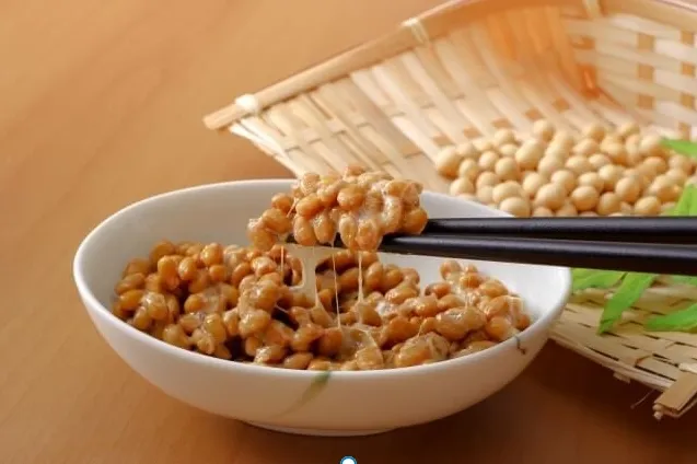 白い皿に盛った納豆を箸で持ち上げ、糸引きの粘り気が見えるクローズアップ。背景に木のザルに納豆豆と緑の葉が並ぶ伝統的な日本の食品イメージ。腎臓に良い納豆の魅力を表す。