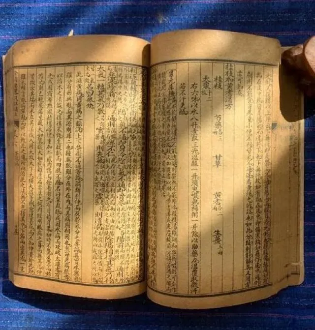 青い布の上に置かれた古い医学書が開かれ、光が当たって乳がん(乳巌)の古代記録を象徴するイメージ
