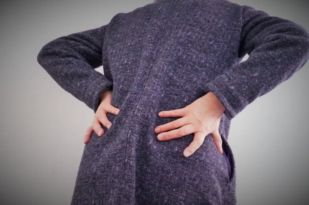 紺色のセーターを着たシニア女性が、腰とお尻の痛みを訴えるように両手で触っている様子。お尻の痛みや坐骨神経痛の症状を表すイラストレーション。