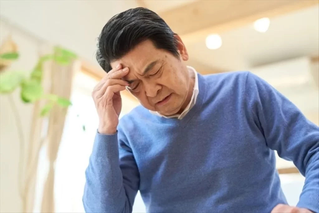 頭を押さえて目を閉じる高齢男性の写真。認知症の不安や混乱を表すイメージ。