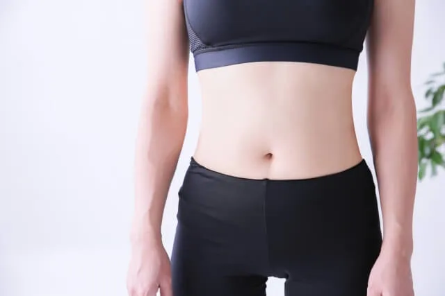 黒いスポーツウェアを着たスリムな女性がお腹を露出して自信たっぷりにポーズ。体脂肪率を下げるダイエット成功のイメージ。