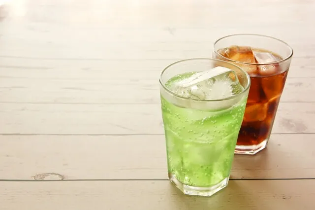 透明なコップに入った緑色の清涼飲料水とコーラ色の清涼飲料水が並んでいる画像。リン酸塩の危険性を示す記事のアイキャッチ。
