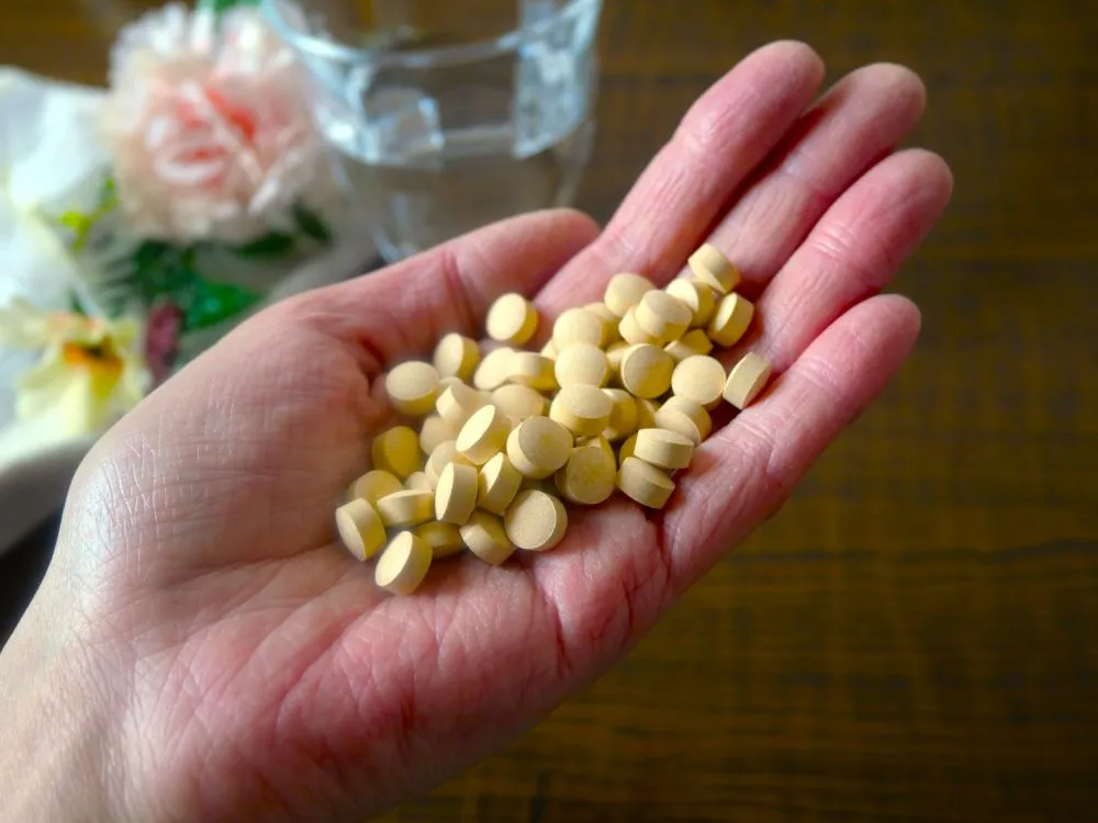 手のひらに黄色い錠剤が山積みになった画像。背景に水の入ったコップと花が配置されている。