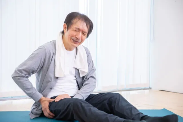 床に座った70代くらいの男性が首にタオルをかけ、右太ももを押さえて痛みに顔を歪めている写真