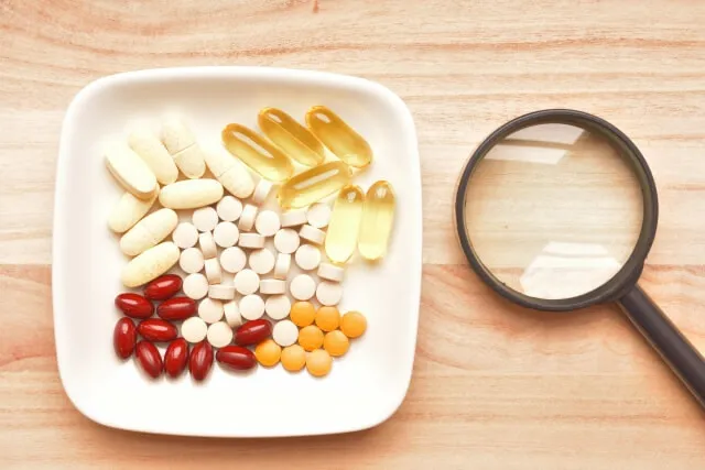 白い皿に並べられた色とりどりのサプリメントのカプセルと錠剤、隣に拡大鏡が置かれた俯瞰写真