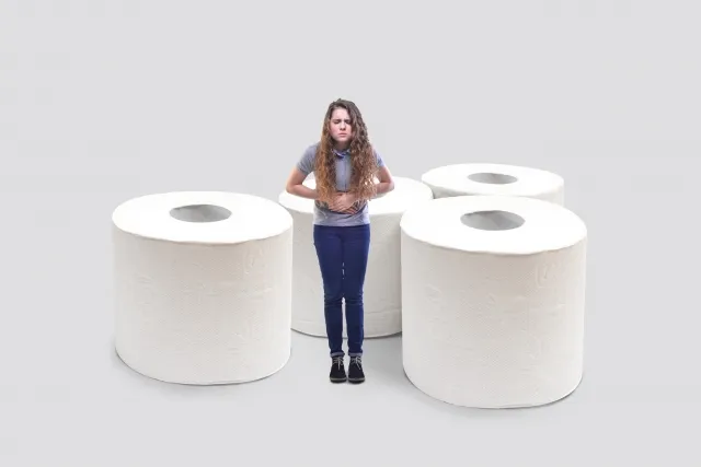 便秘に苦しむ金髪の女性が大きなトイレットペーパーの前に立ち、お腹を抱えて辛そうにするイラスト。1回の足つぼ整体で改善した症例を象徴。
