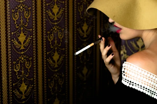 大きな帽子をかぶった白人女性が赤い口紅でタバコを吸い煙を吐く横顔。ストレスと喫煙が肺がんを悪化させた患者のイメージ。