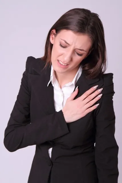 スーツ姿の白人女性が心臓の痛みで目を閉じ、右手で胸を強く押さえている様子。心臓発作の症状を表現した画像。