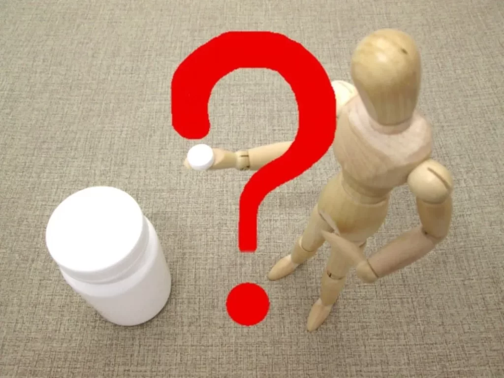 木の人形が右手に薬を持ち、前の白い薬瓶と赤い疑問符が薬の危険性を示している画像
