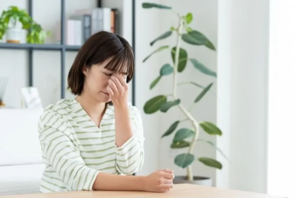 シマシマの七分袖を着た女性がソファーに座り、左手で眉間を押さえて疲れた表情をしている様子