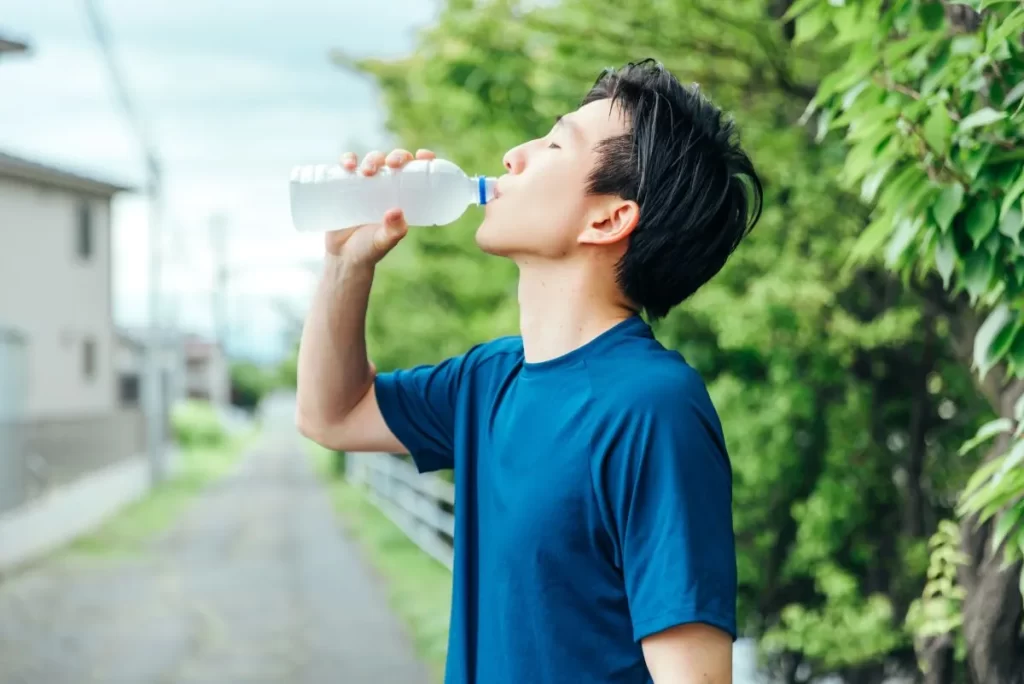 青い半袖の男性が頭を濡らしながらペットボトルの水を飲んで水分補給している様子。背景に家と緑の葉が見える。