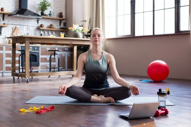 ヨガマットに座り瞑想する白人女性。周囲にバランスボール、ノートPC、お茶があり、後ろにテーブルと本棚が見える