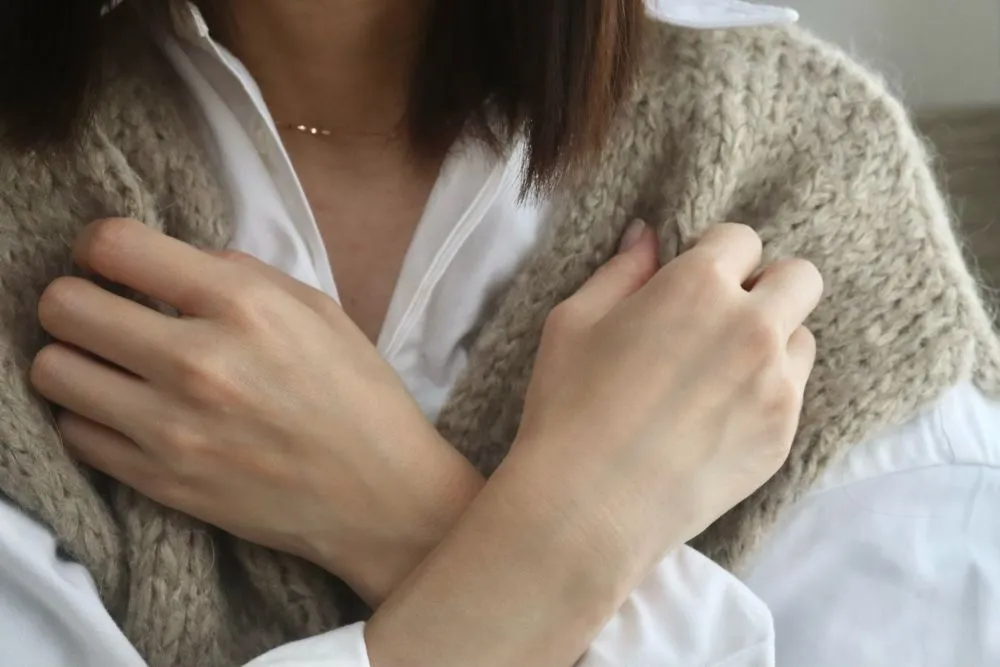 ニットベストを着た女性が寒さに震えながら両手で肩を抱える様子。手足の冷え性を象徴するイラストで、中医学の未病予防を促す。