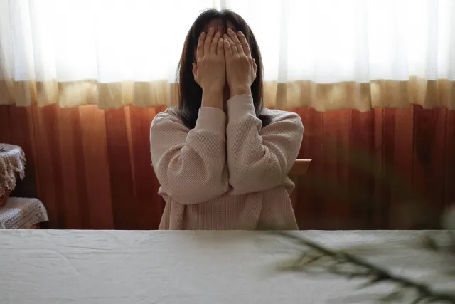 カーテンの前で両手で顔を覆い、不安そうに座る女性。