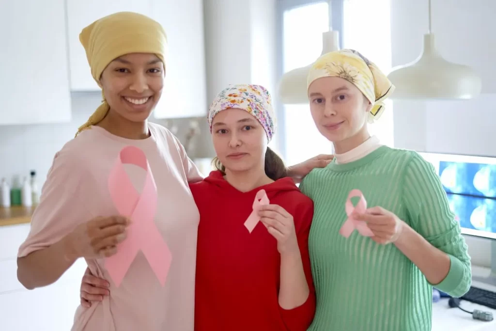 頭にスカーフを巻いた黒人、白人、アジア人の3人の女性が肩を並べ、片手にピンクリボンを持ち、乳がん意識向上を象徴する多様な連帯のイメージ