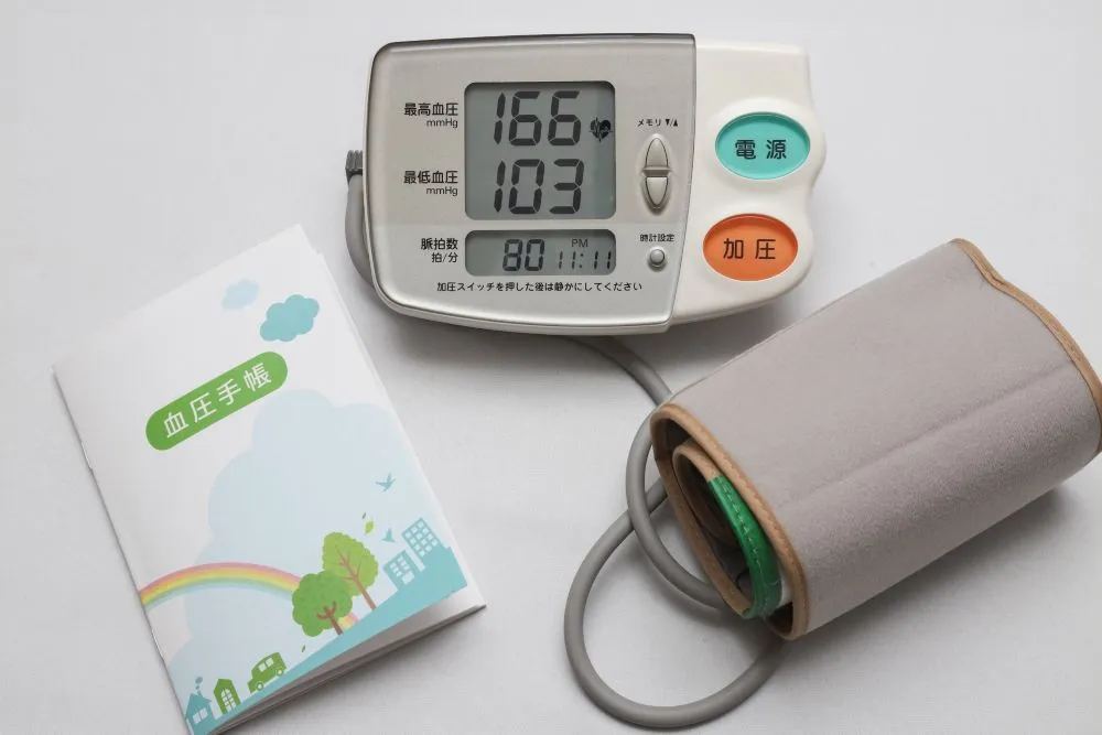 血圧計と血圧手帳が木製の机の上に置かれている画像。血圧手帳には血圧の記録が書かれている。