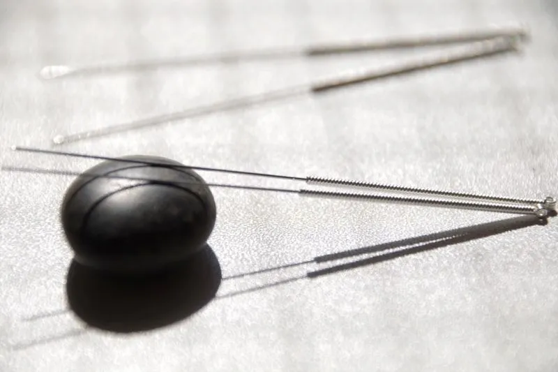 黒い丸い石の上に針先を乗せた2本の鍼、背景に薄く浮かぶ鍼のシルエット。鍼灸のシンプルな道具を象徴するイメージ。
