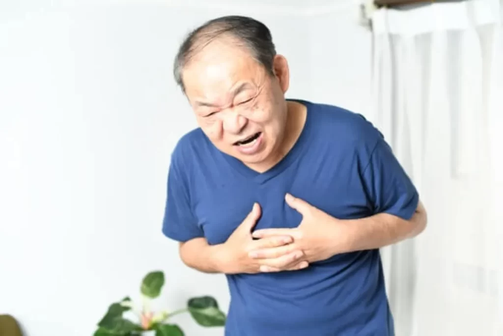 青い半袖を着た81歳の高齢男性が胸を両手で押さえ、心臓の痛みに苦しむ表情をしている