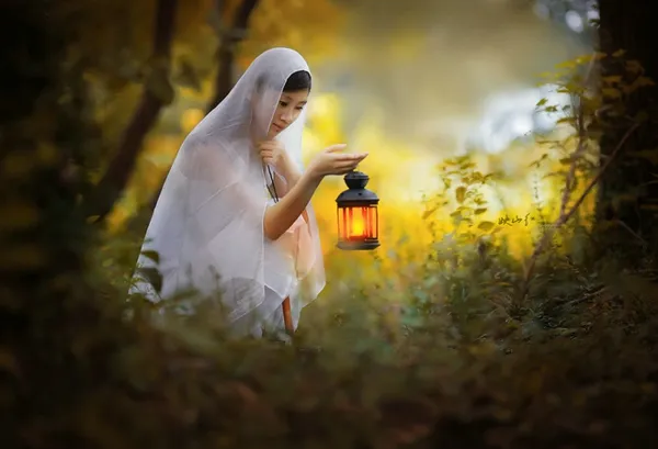 絹のスカーフを頭に被った女性が、暗い森の中で提灯の灯りを頼りに何かを探す神秘的な姿