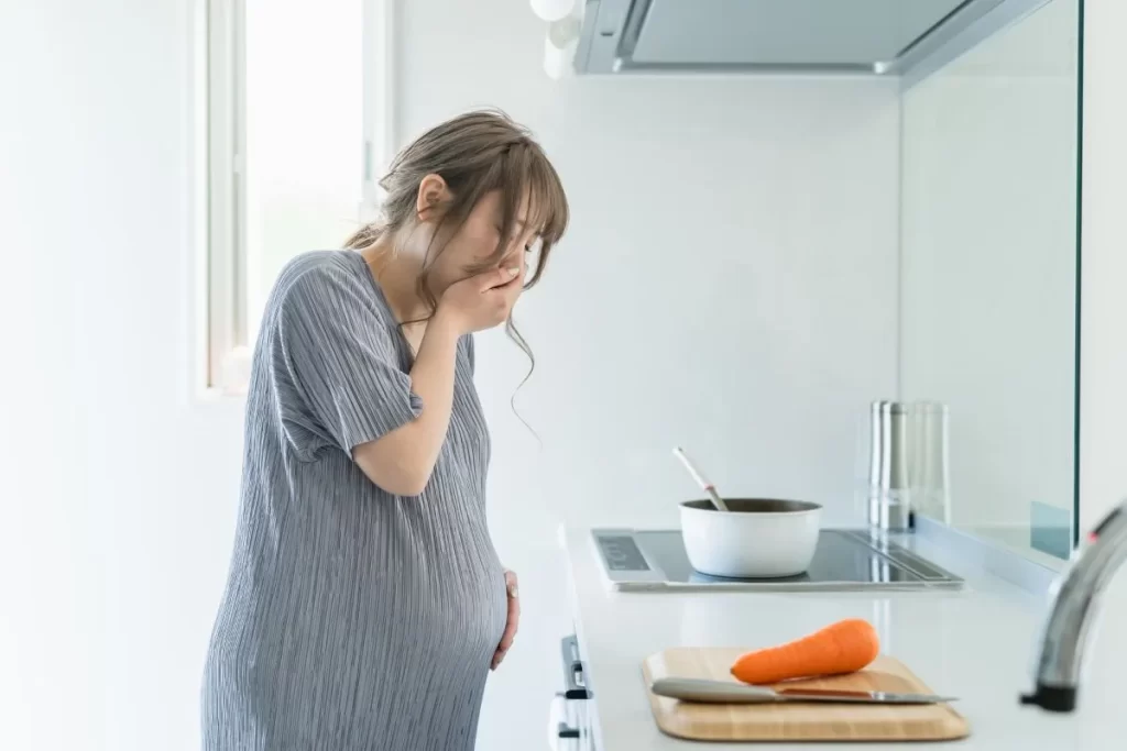 妊娠中の女性がつわりで吐き気を抑えながらキッチンに立つ様子、まな板と野菜の前で口を押さえている