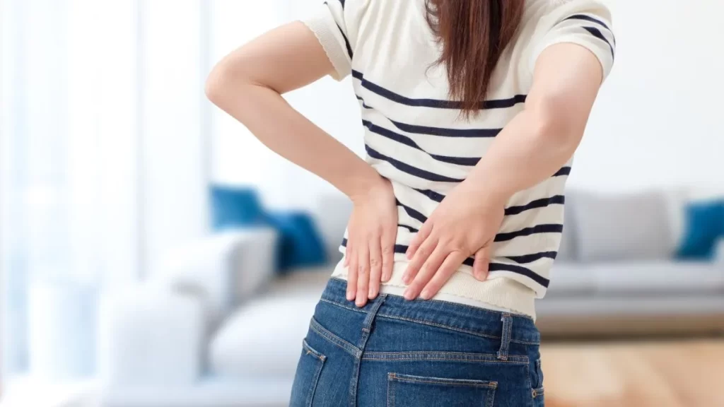 ジーパンとシマシマの半袖セーターの女性が、両手で腰を押さえて痛みを感じている様子