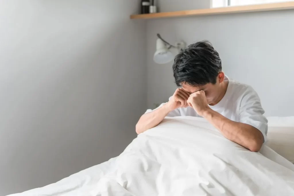 寝不足で目を押さえる男性が白い寝具の中で起き上がっている様子。睡眠障害と倦怠感のイメージ。