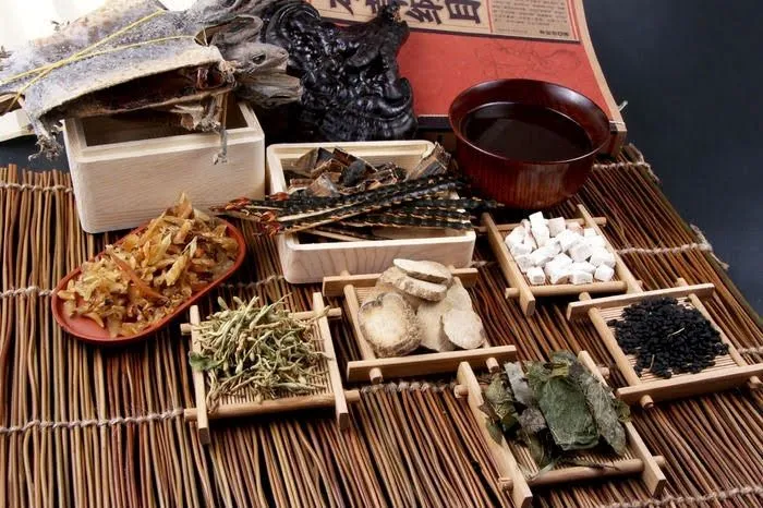 木のザルに並ぶ漢方生薬と木容器の漢方薬汁、本草綱目書籍の文字が写る中医学治療のイメージ