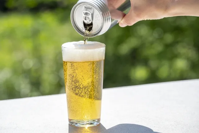 缶ビールをグラスに注ぐ様子のクローズアップ写真。背景に緑の葉がぼんやり映る