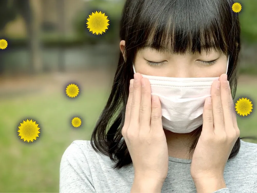 マスクをして目を閉じる花粉症に苦しむ女性の横顔。周囲に黄色いトゲトゲの花粉が飛んでいる