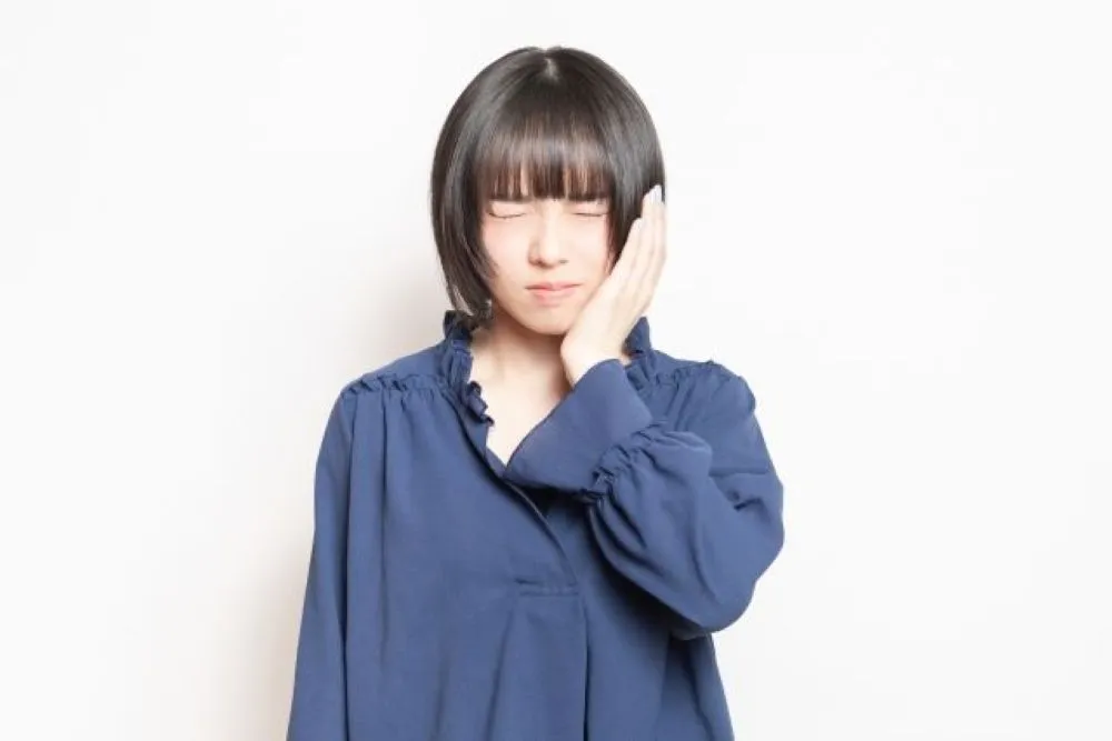 歯の痛みで左頬を押さえる女性（紺色の長袖を着用）