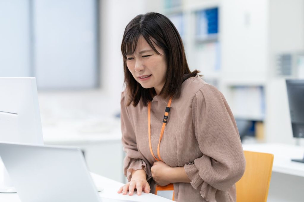 リーキーガット症候群で腹痛に悩む女性会社員。パソコン作業中に左手でお腹を押さえている様子