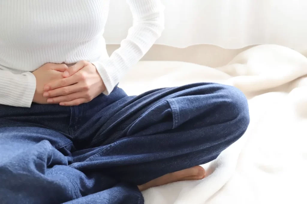 下腹部の痛みを抱える女性があぐらで座り、両手で下腹を押さえている様子（子宮がんの不調をイメージ）
