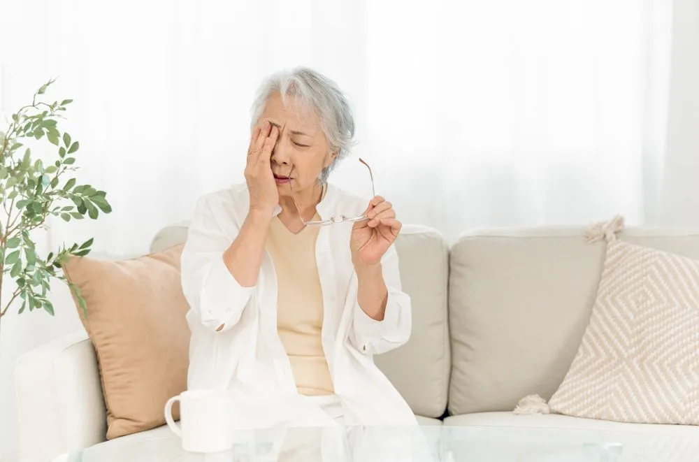 白内障の症状で右目を触る白髪の66歳女性、ソファーに座り左手にメガネを持つ様子