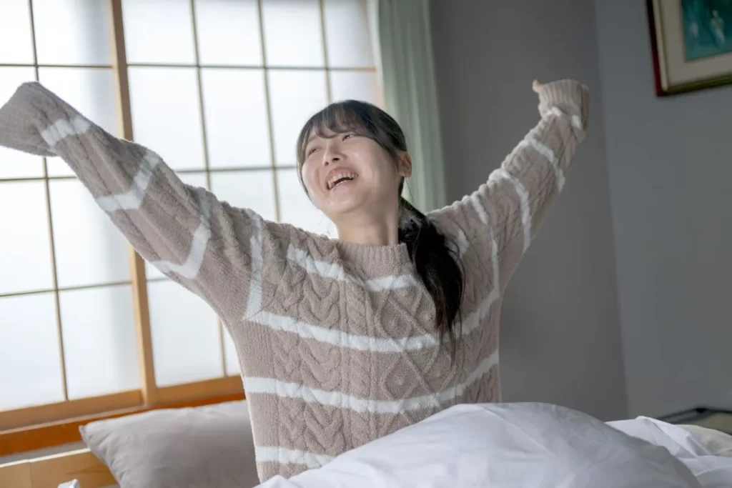 白とグレーのセーターを着た女性が布団から起き上がり、両手を上げて満足そうに笑っている。後ろには和風の木製ガラス戸がある。