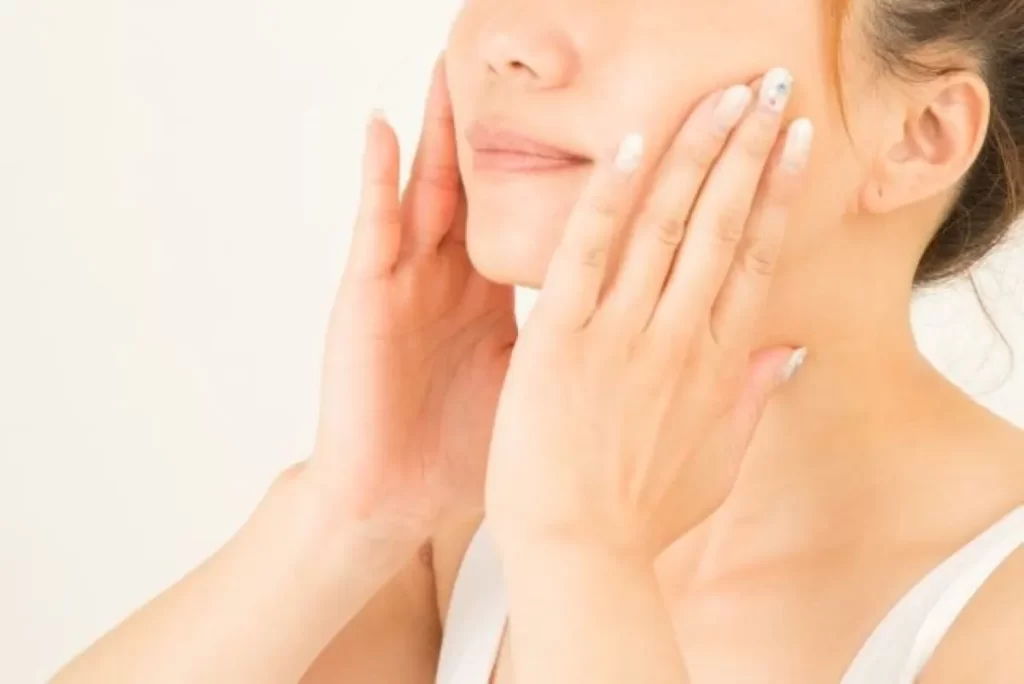 女性が両手で頬を優しく触り、綺麗な肌を確かめる様子（白いタンクトップ着用）