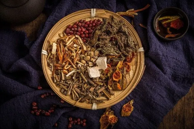 紺色のシルク生地の上に藤編みの籠で並べられたさまざまな漢方生薬。周りに散らばった薬材と黒い陶器が見える。