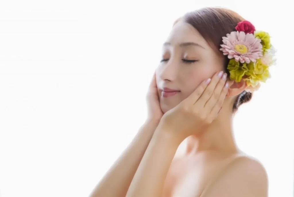 足つぼと鍼灸治療で肌の調子が良くなった女性が目を閉じて顔を優しく触っている様子。左側に美しい花が飾られている