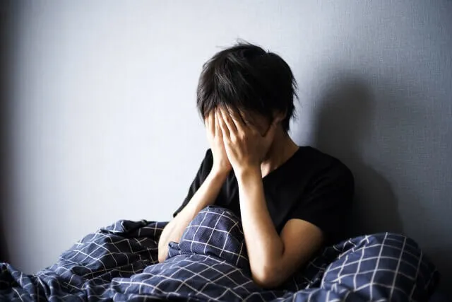 不眠症に悩む男性がベッドに座り、両手で顔を覆って苦しむ様子。寝汗や冷え性の症状改善を求める漢方症例のイメージ。