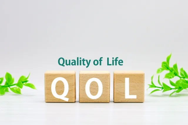 QOL（Quality of Life）の文字が書かれたブロックと緑の葉、背景に青い「Quality of Life」の文字。生活の質を象徴する画像