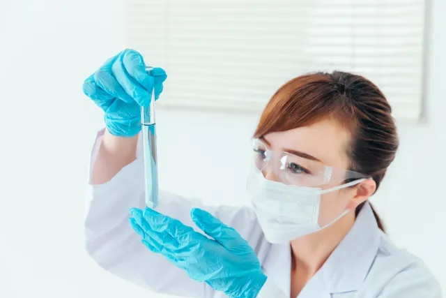 青い手袋とマスクをした女性科学者が試験管を覗き込む画像。製薬会社が力を入れて開発する新薬の研究シーン