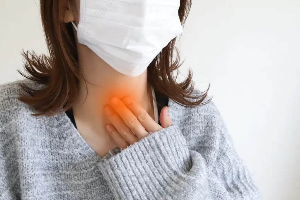 喉の痛みで左手を当てているマスク姿の女性。扁桃腺炎を示すように喉が赤くなっている。