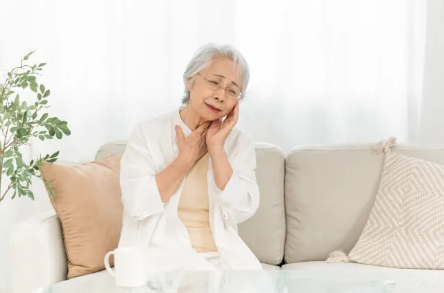 白髪の高齢女性が白い服を着てソファに座り、眉間に皺を寄せて喉の痛みを気にする様子。喉がん術後の呼吸困難や咳の症状を表すイメージ。