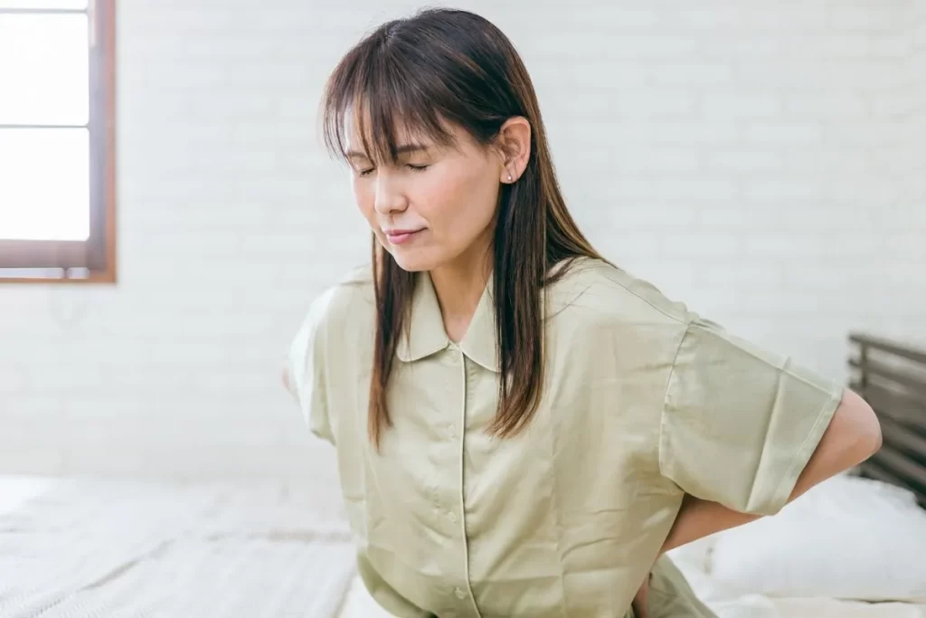 パジャマ姿のシニア女性がベッドに座り、両目を閉じ、両手で腰を押さえている画像