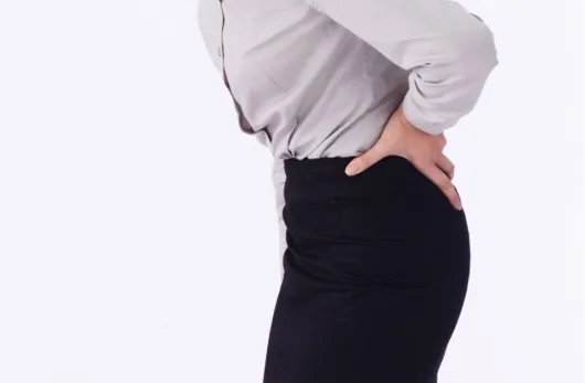 妊娠中の女性が坐骨神経痛でお尻を気にする様子。黒いスカートとグレーのワイシャツを着て、痛みを和らげようと触っている。