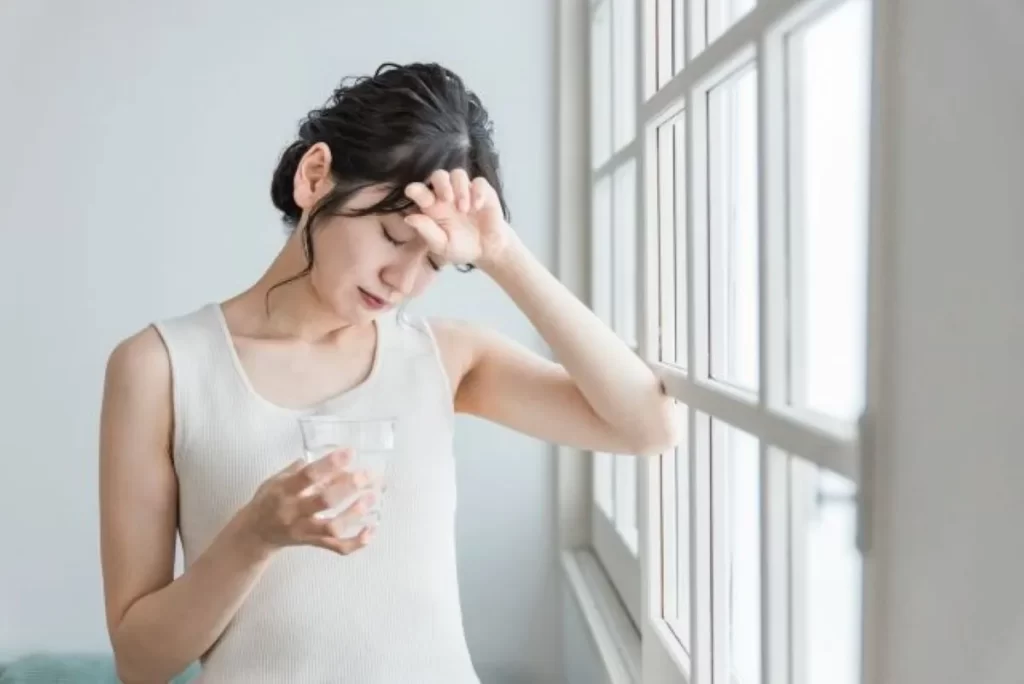 白いタンクトップの女性が水のコップを持ちながら額に手を当て、うつ病や頭痛のつらさを感じている様子。