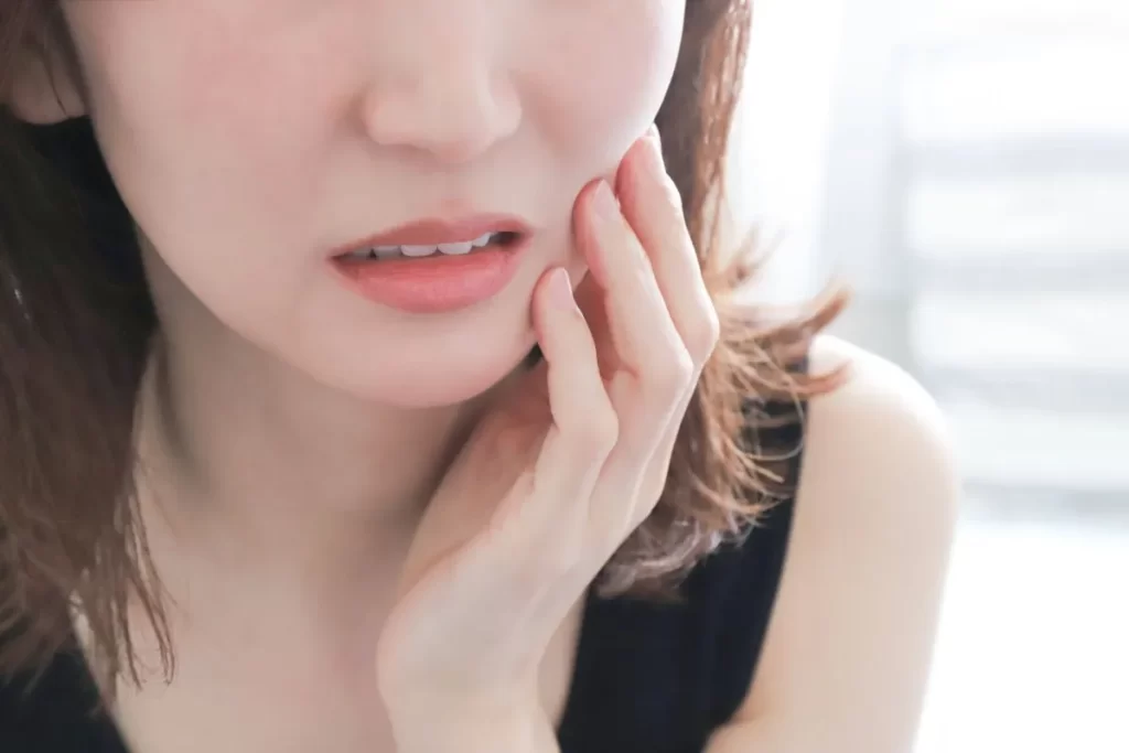 冷たい水を飲んで歯がしみるため、左頬を押さえて痛そうな表情をする女性