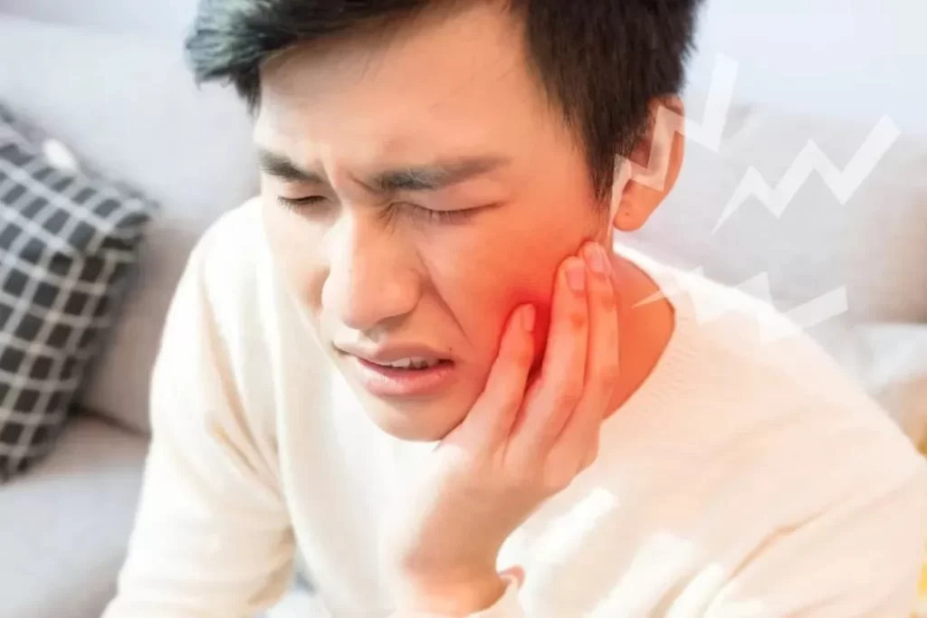 白いセーターを着た男性がソファーに座り、左ほっぺの痛みで顔をゆがめながら手で触れているイラスト。頬の周囲に電撃のような痛みを示す雷マークが描かれている。