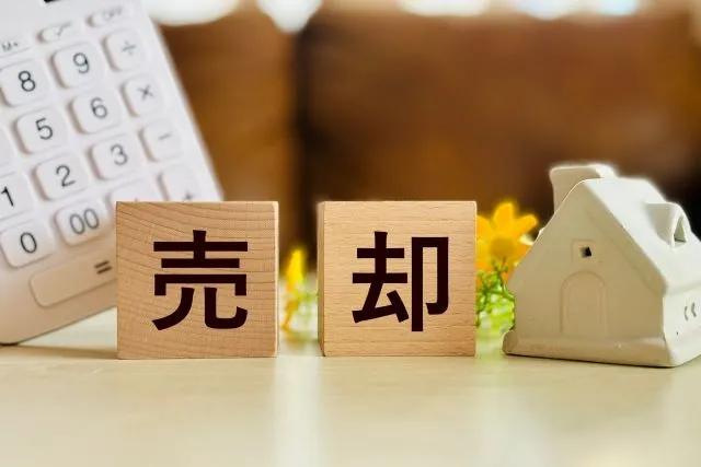大きな「売却」の文字が前景にあり、背景に電卓やペンがうっすら見える画像。病気の治療費で自宅マンションを売却せざるを得ない状況を象徴。