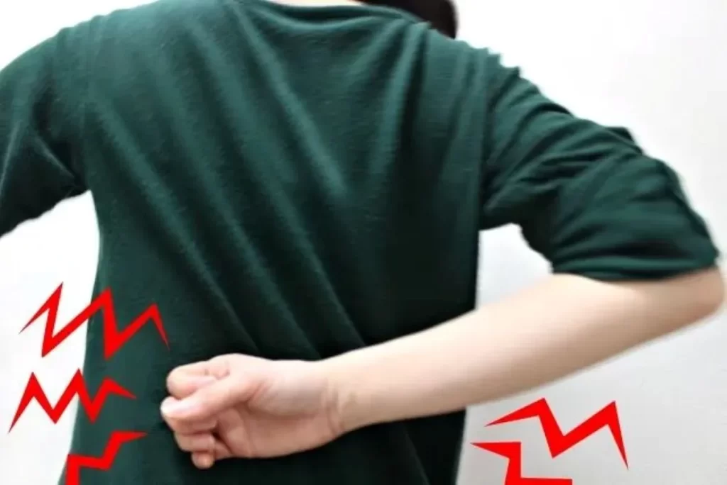 黄緑色の長袖を着た女性が腰を叩く背中姿。腰回りに赤い電撃のような痛みがあり、子宮内膜症や術後後遺症の激しい腰痛を象徴。