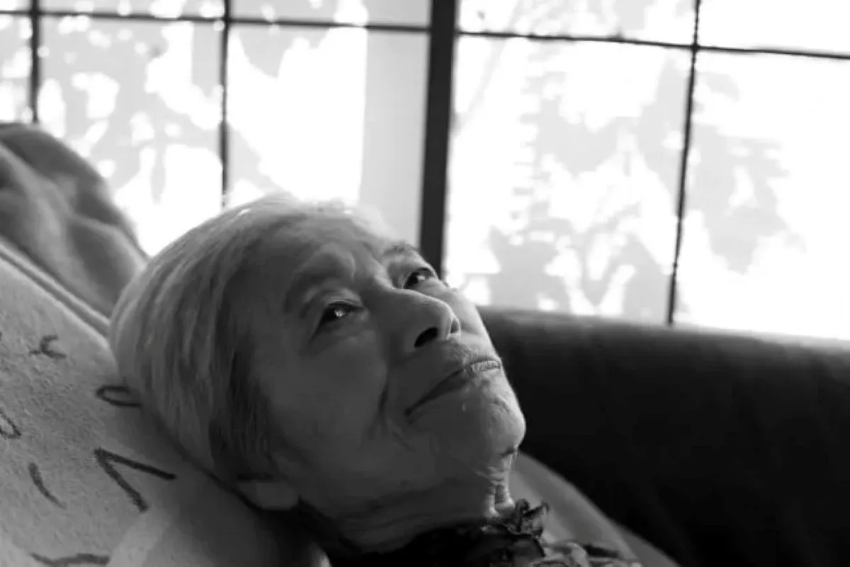 寝たきりでベッドに横たわる白髪の高齢女性が、静かに天井を見つめている白黒写真。認知症と薬害による長期介護の現実を象徴する。